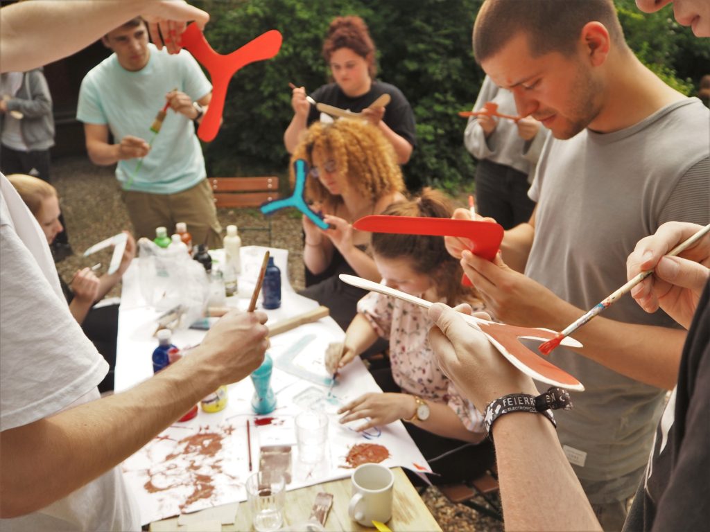 junge Menschen sitzen und stehen um Tisch im Freien und bemalen Bumerangs