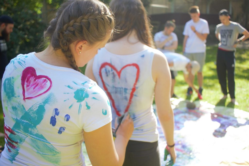 Seminargruppe, draußen, im Vordergrund junge Frau, die das T-Shirt einer anderen jungen Frau bemalt