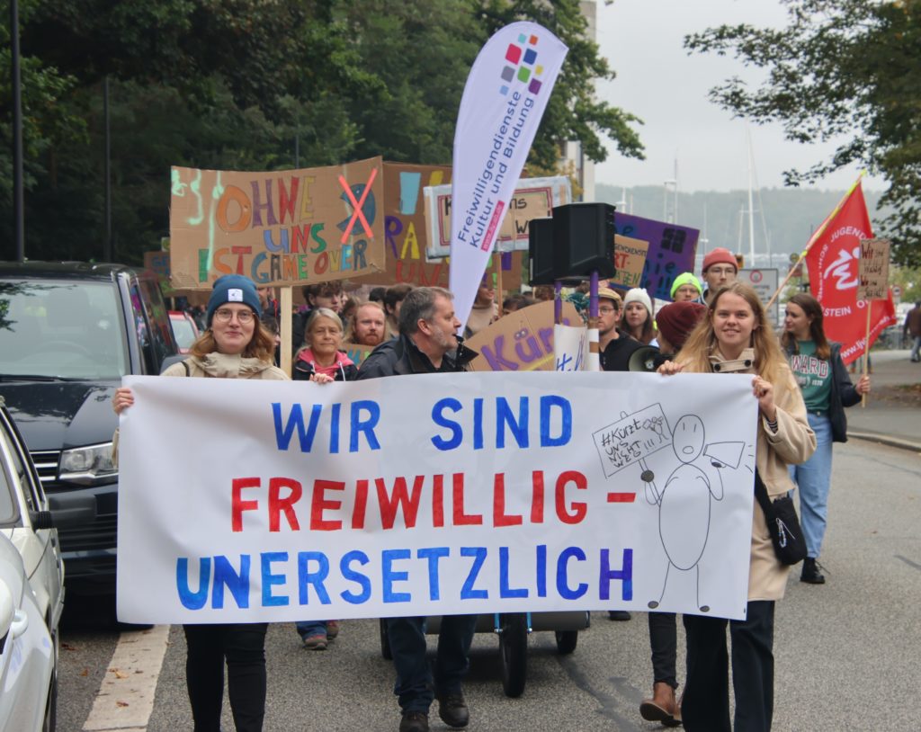 Demonstrationszug, im Vordergrund großese Banner mit der Aufschrift: "Wir sind Freiwillig - unersetzlich"